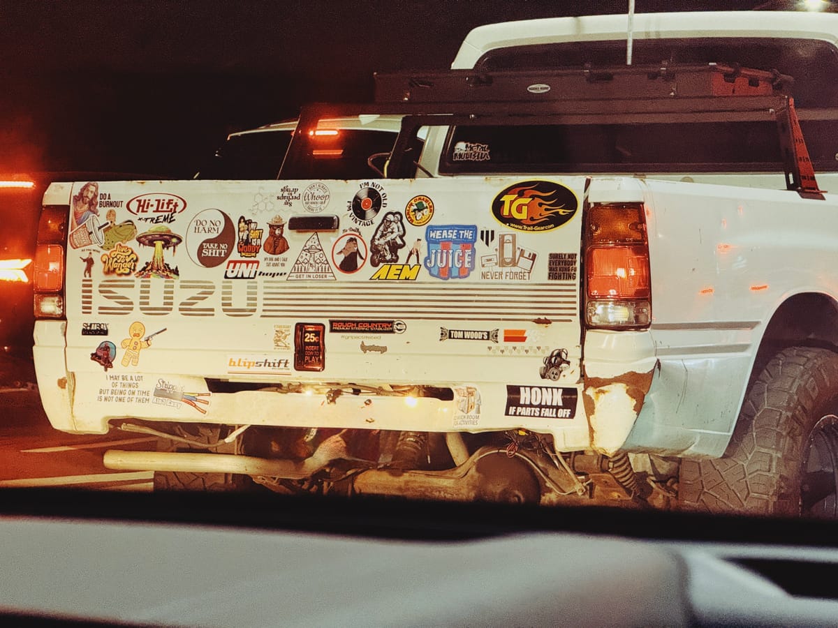 Telephoto image of the back of a white pickup truck with many humorous bumper stickers all over its tailgate, like a yin-yang design that says, "Do no harm, take no shit"
