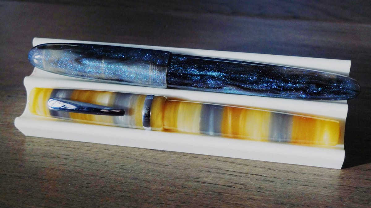2 fountain pens sitting in a dual pen stand: a blue-green-black shimmery, cigar-shaped pen in the top slot; a yellow, honey, and gray striped pen with clip in bottom slot