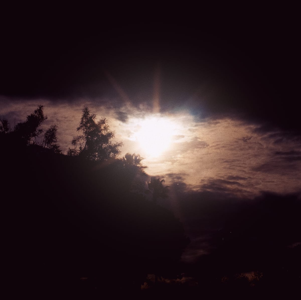 High-contrast scene of sun shining bright, through cloud cover with dark tree silhouettes covering the bottom 2/3 of the frame, and dark clouds in top 1/4
