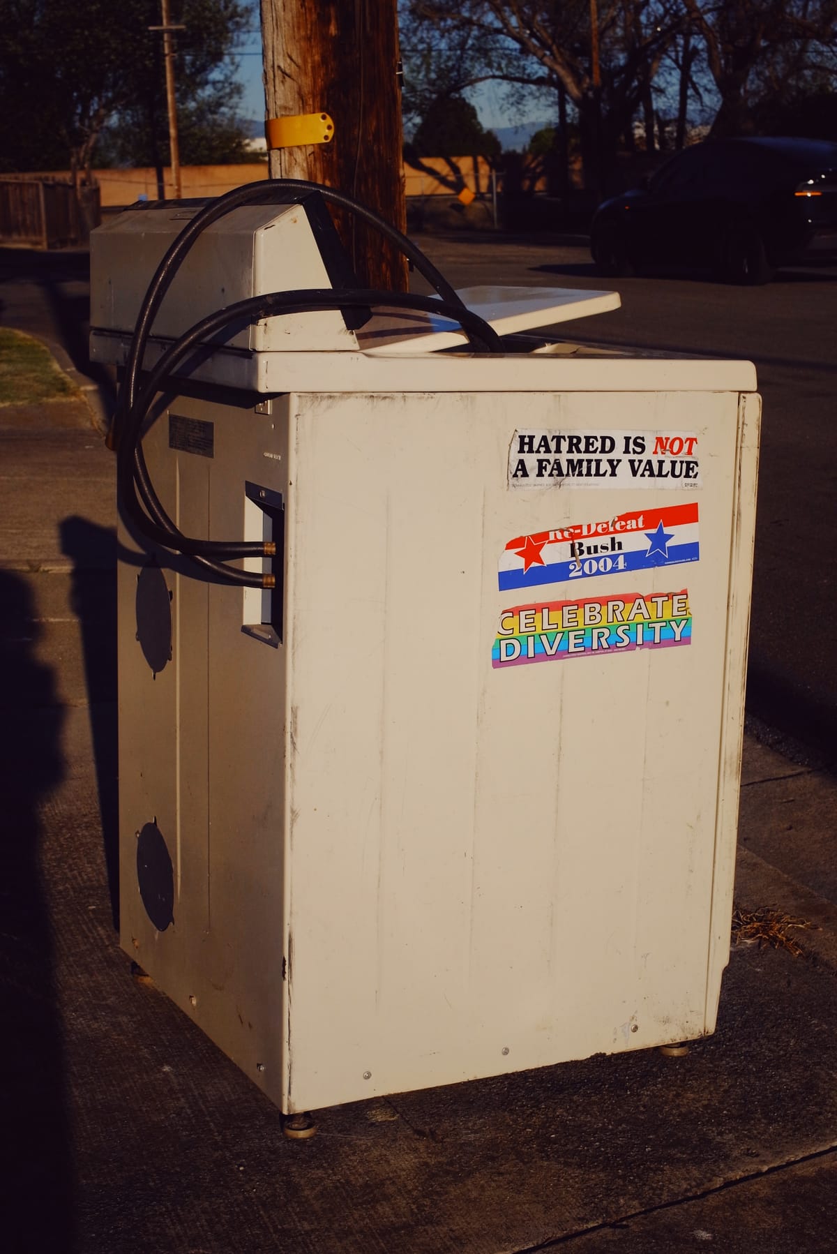 Appliance sitting on a sidewalk with some bumper stickers on its side: "Hatred is not a family value", "Re-Defeat Bush 2004", and "Celebrate Diversity (with a rainbow background)"