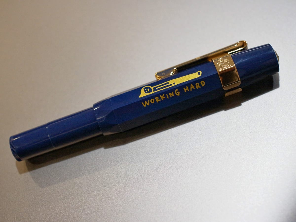 Medium-dark blue Kaweco Sport fountain pen with a gold clip and a painted graphic of a person lying flat on the ground, captioned "working hard" underneath
